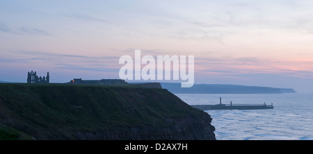 Whitby Abbey ruins at sunset, silhouetted & perched high on coastal clifftop headland, overlooking pier & sea - scenic Yorkshire Coast, England, UK. Stock Photo