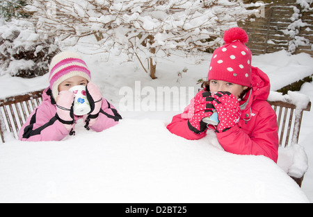 Two young girls have a hot drink sitting on snow covered garden furniture during winter in the south of England Stock Photo