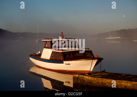 White boat in a quiet harbor in the middle of a foggy morning with the moon on the horizon Stock Photo