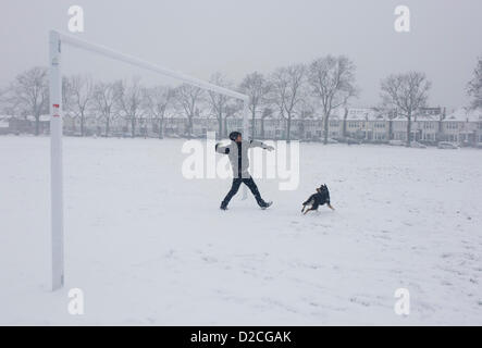 A pet owner is about to throw a yellow tennis ball for his Alsatian dog during a snowy day in south London park. During a prolonged cold spell of bad weather, snow fell continuously on the capital on Sunday, allowing families the chance to enjoy the bleak conditions, here in Ruskin Park in the borough of Lambeth. Pulling back his right arm, the man pauses before pitching the ball away from under a football goalpost, part of a soccer pitch used by local teams. The dog waits for the ball's trajectory before tearing off into the distance near Edwardian period houses beneath 100 year-old ash trees Stock Photo
