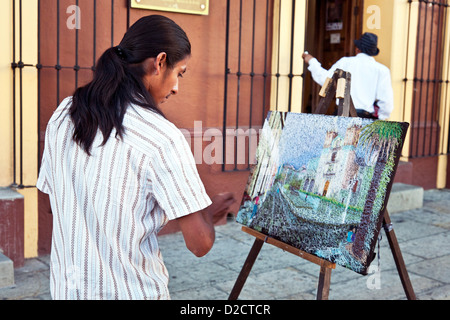 young Oaxacan artist at easel in street pulls silk thread through his impressionistic woven landscape of Santo Domingo Church Stock Photo
