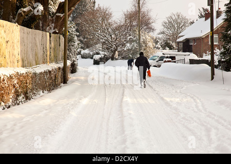 A man walks along a snow covered road in Devon, UK with another person clearing snow with a shovel in the background. Stock Photo