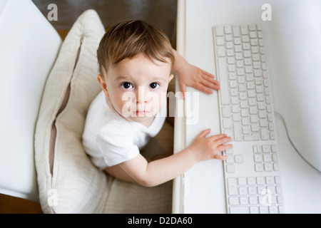 A toddler sitting at a desk with a computer keyboard Stock Photo