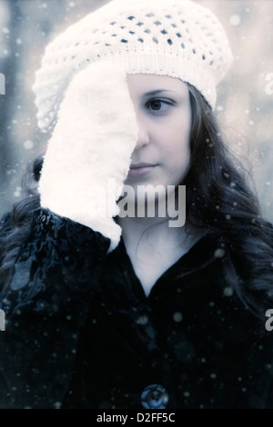 a woman in a black coat with a white cap is holding a gloved hand in front of one of her eyes Stock Photo