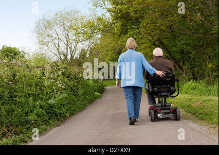 Mature couple walking - married older retired couple in the countryside while one drives a motorised mobility scooter, UK Stock Photo