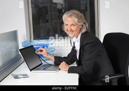 Portrait of senior businesswoman examining laptop with the use of stethoscope at office desk Stock Photo
