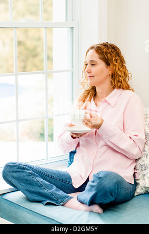 Smiling caucasian woman relaxing on couch by window holding cup of coffee Stock Photo