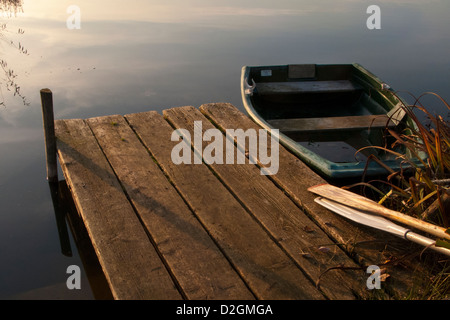 Wooden Jetty pier by lakeside in late afternoon light with rowing boat and wood oars Stock Photo