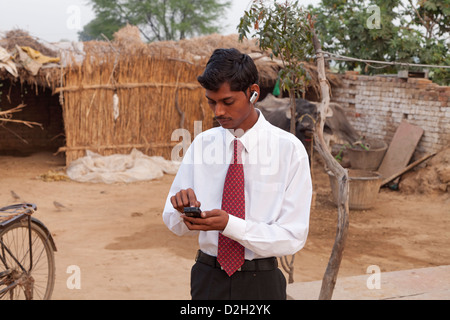 India,Uttar Pradesh, Agra, Young adult man wearing shirt and tie using smartphone and bluetooth headset outside village home Stock Photo