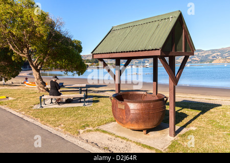 A try pot, relic of Akaroa's days as a whaling station, on the waterfront of the harbour. Woman sitting on bench usig iPad. Stock Photo