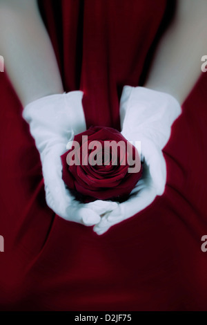 a woman in a red dress with white gloves is holding a red rose on her lap Stock Photo