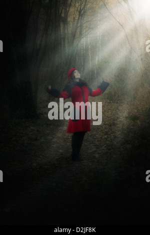 a woman in a red coat is standing in a dark forest in a spot of sunlight Stock Photo