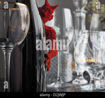a little red devil, modelling clay, peeks out from behind a bottle of wine. Some glasses are spread out on the inlaid table. Stock Photo