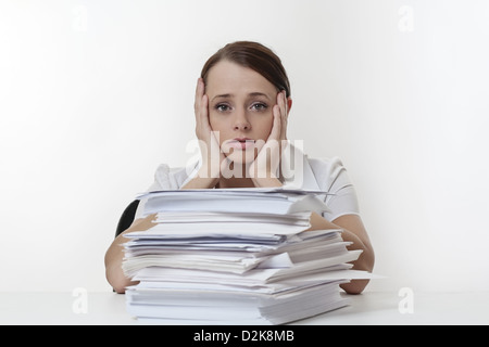 A stressed female with her head in her hands, sitting at her desk with a large pile of paper work stacked in front of her.  Stock Photo
