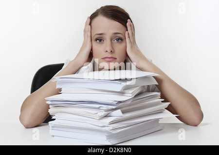 A stressed female with her head in her hands, sitting at her desk with a large pile of paper work stacked in front of her.  Stock Photo