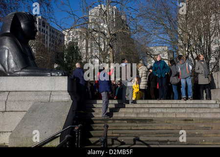 A tour group organised by WalkLondon stop by a sphinx to hear about the ancient Egyptian obelisk known as Cleopatra's Needle, on the Embankment WC2. Cleopatra's Needle is flanked by two faux-Egyptian sphinxes cast from bronze that bear hieroglyphic inscriptions that say netjer nefer men-kheper-re di ankh (the good god, Thuthmosis III given life). These Sphinxes appear to be looking at the Needle rather than guarding it. This is due to the Sphinxes' improper or backwards installation. Stock Photo