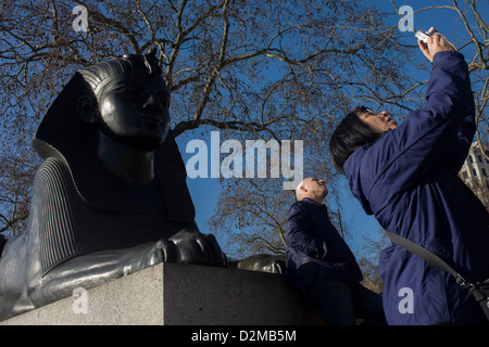 A tour group organised by WalkLondon stop by a sphinx to hear about the ancient Egyptian obelisk known as Cleopatra's Needle, on the Embankment WC2. Cleopatra's Needle is flanked by two faux-Egyptian sphinxes cast from bronze that bear hieroglyphic inscriptions that say netjer nefer men-kheper-re di ankh (the good god, Thuthmosis III given life). These Sphinxes appear to be looking at the Needle rather than guarding it. This is due to the Sphinxes' improper or backwards installation. Stock Photo