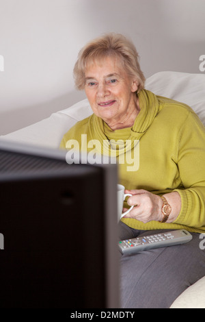 Mature lady sitting holding the a mug of tea or coffee, watching TV. Stock Photo