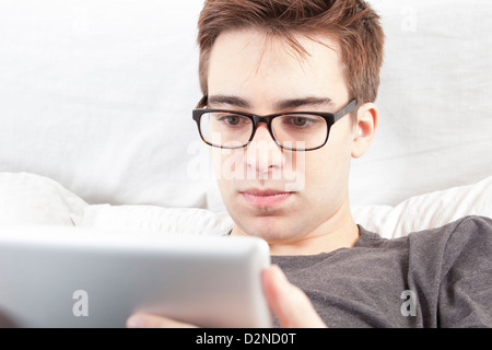 Young male wearing glasses looking at the screen of his digital tablet, sitting on his bed. Stock Photo
