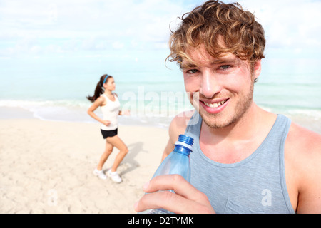 Portrait of fit young man drinking water with woman jogging in background on beach Stock Photo