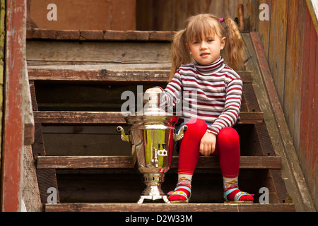 Funny lovely little girl posing sitting near the Russian Samovar on the porch of the farmhouse. Stock Photo
