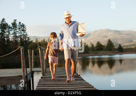 Smiling grandfather and grandson with toy sailboat holding hands and walking along dock over lake Stock Photo