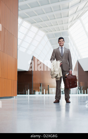 Portrait of smiling businessman holding coat and briefcase in lobby Stock Photo