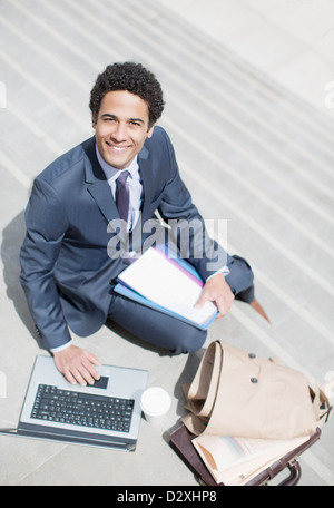 Portrait of smiling businessman with paperwork using laptop on sunny stairs Stock Photo