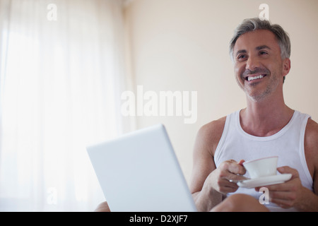 Smiling man drinking coffee and using laptop in bedroom Stock Photo