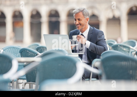 Smiling businessman drinking coffee and using laptop at sidewalk cafe Stock Photo