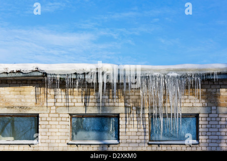 Very cold in winter concept. Huge icicles hanging on roof of a brick old rustic building on a background of frozen blue sky. Sun Stock Photo
