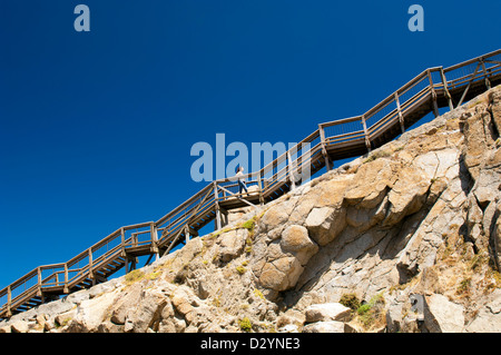 Young woman climbs up steep stairs on a mountain with blue sky behind as copy space Stock Photo