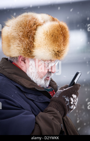 Senior man wearing fur hat and using smart phone in snowstorm Stock Photo