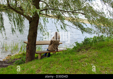 young blond woman sit on wooden bench under willow tree branches and admire lake landscape. Stock Photo