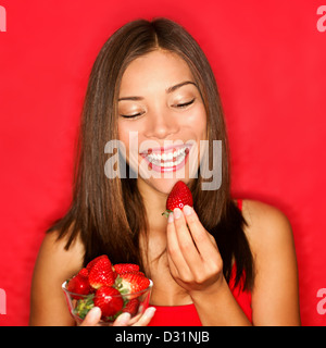 Smiling young mixed race Chinese Asian / Caucasian woman eating strawberry on red background Stock Photo