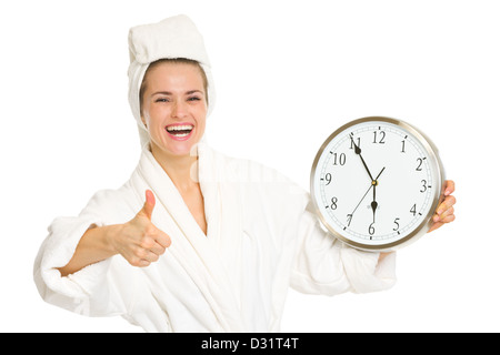 Smiling young woman in bathrobe showing clock and thumbs up Stock Photo
