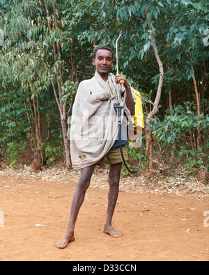 A young barefoot boy carrying a pesticide sprayer and backpack on a dirt road Stock Photo