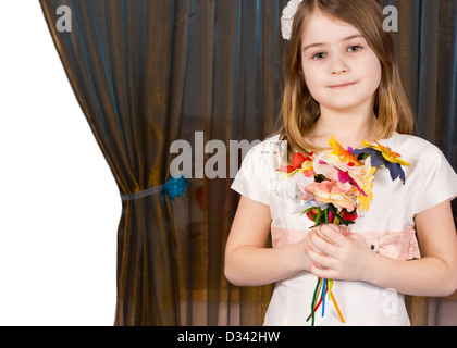 Beautiful little girl posing with a handful of colourful artificial flowers against a curtain drawn aside on the left to reveal Stock Photo