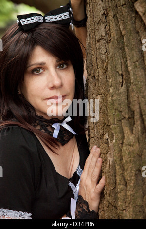 Cute white woman in a maid's outfit, standing outside against the rough texture of a tree and looking at the camera Stock Photo