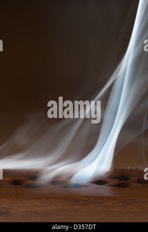 Burning incense smoke drifting whirling into a darkened room from exit holes in an enclosed wooden holder Stock Photo