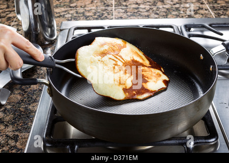 Horizontal photo of main focus on pancake being flipped in hot frying pan on top of gas stove range Stock Photo