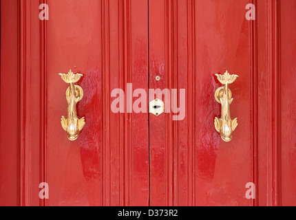 Two brass door knockers in the shape of fish on a bright red door. Stock Photo