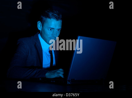 A suspicious looking man in a business suit working on a laptop computer in the dark. Could be a hacker, or government snooper Stock Photo