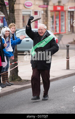 Elderly male competitor (smiling & raising his hat to crowd) taking part in traditional, Ilkley Rotary Pancake Race - The Grove, Ilkley, West Yorkshire, UK. Stock Photo