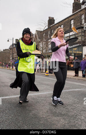 Female competitors (flipping pancakes) taking part, running in traditional, annual Ilkley Rotary Pancake Race - The Grove, Ilkley, West Yorkshire, UK. Stock Photo