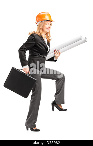 Full length portrait of a mature female architect with helmet holding blueprints and suitcase, walking with big steps isolated o Stock Photo