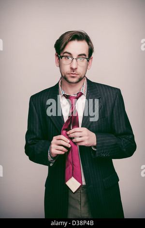 Businessman in his 20s wearing pinstripe suit jacket, white shirt, red tie and glasses looks to camera while doing up his tie. Stock Photo