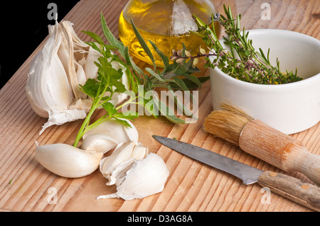 Garlic, parsley, sage, rosemary and thyme with extra virgen olive oil in a jug, a brush and knife. on an antique chopping board. Stock Photo