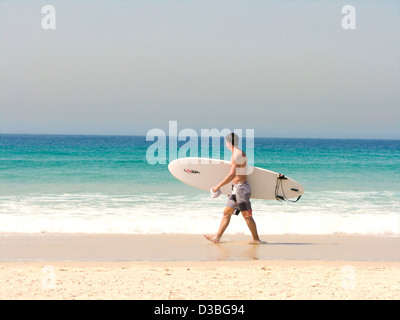 A lone surfer walks along Bondi Beach carrying his surfboard looking at the waves. Stock Photo