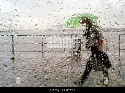 (dpa) - Seen through the rain drops on a window pane a woman carrying a green umbrella walks past a street cafe in the harbour in Hamburg, 4 July 2003.